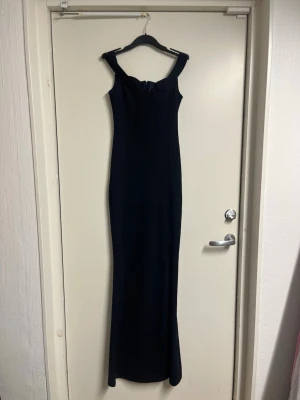 Marinblå offshoulder långklänning - Elegant marinblå långklänning med offshoulder-design och figurnära passform. Klänningen har en stilren och enkel look som framhäver siluetten. Perfekt för dig som vill ha en klassisk och tidlös stil. Materialet känns mjukt och följsamt mot kroppen.