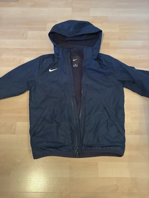 Mörkblå Nike vindjacka med huva - Snygg mörkblå vindjacka från Nike med huva och dragkedja framtill. Jackan har två sidofickor och en vit Nike-logga på bröstet. Tillverkad i lätt och tåligt syntetmaterial, perfekt för blåsiga dagar.