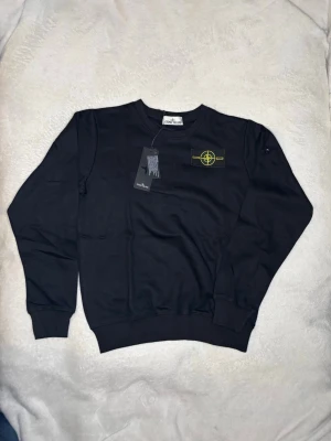 Svart sweatshirt från Stone Island - Cool svart sweatshirt från Stone Island med det klassiska kompassmärket i gult och grönt på bröstet. Tröjan har rund hals, ribbade muddar och är tillverkad i mjukt bomullsmaterial. Perfekt för dig som gillar streetwear och vill ha något stilrent.