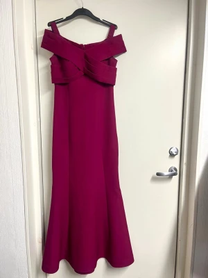 Vinröd långklänning med offshoulder - Vinröd långklänning med elegant offshoulder-design och breda band som korsas framtill. Klänningen har figurnära passform och är tillverkad i ett slätt material som ger en lyxig känsla. 