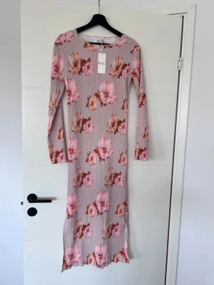 Blommig långklänning från BYIC - Långklänning från BYIC med blommigt mönster i rosa och bruna toner på en ljusbeige bakgrund. Klänningen har lång ärm och rund halsringning. Materialet är stretchigt och lätt, perfekt för vår och sommar.