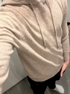 Beige stickad hoodie med huva - Mysig beige stickad hoodie med huva och dragsko. Tröjan har ribbade muddar vid ärmslut och nederkant. Perfekt för en avslappnad och stilren look. Materialet känns mjukt och skönt mot huden.
