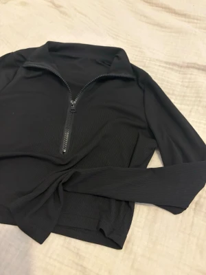 Svart ribbad polotopp med half zip - Snygg svart ribbad polotopp med lång ärm och half zip-dragkedja framtill. Toppen har en croppad passform och är tillverkad i ett stretchigt material som sitter bekvämt. Perfekt till jeans eller kjol för en trendig look. Använts en gång och sedan samlat dam i garderoben. Kontakta mig vid intresse eller egna bud. Finns i Norrköping 