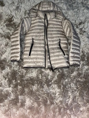 Ljus dunjacka med huva McKinley - Snygg ljus dunjacka från McKinley med huva och svarta dragkedjor på fickorna. Jackan har quiltad design och är lätt vadderad, perfekt för kalla vinterdagar. Passar dig som vill ha en stilren och funktionell jacka med sportig känsla.