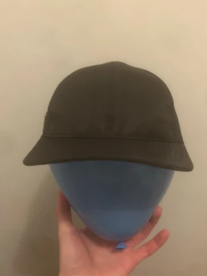 Prada nylon keps  - Snygg svart keps från Prada med den ikoniska metalltriangeln framtill där det står 'PRADA MILANO'. Kepsen har klassisk form med böjd skärm och är tillverkad i ett glansigt nylonmaterial som ger en exklusiv känsla.