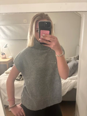 Grå stickad ärmlös polotröja - Säljer en grå stickad ärmlös polotröja med ribbad kant nedtill och vid halsen. Tröjan har en avslappnad passform. Materialet känns mjukt och mysigt. Mycket bra skick