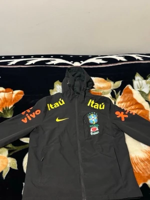 Svart Brasil Nike vindjacka med huva - Snygg svart vindjacka från Nike med Brasiliens landslagslogga och sponsorer i gult och orange. Jackan har huva med dragsko, hel dragkedja och fickor. Perfekt för dig som gillar fotboll och vill ha en sportig stil.