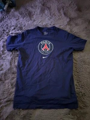 Mörkblå PSG t-shirt från Nike - Snygg mörkblå t-shirt från Nike med stort Paris Saint-Germain-tryck på bröstet och klassisk Nike-logga under. T-shirten har rund hals och korta ärmar. Perfekt för dig som gillar fotboll och vill representera PSG.