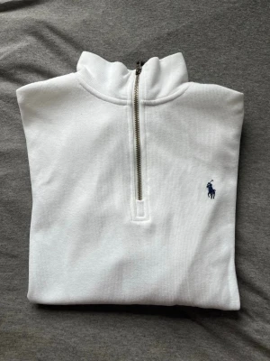 Vit halvzip tröja från Polo Ralph Lauren - Vit långärmad tröja med halvzip och krage från Polo Ralph Lauren. Tröjan har ett diskret broderat blått logomärke på bröstet och är tillverkad i ett mjukt, ribbat material. Perfekt för en clean och stilren look.