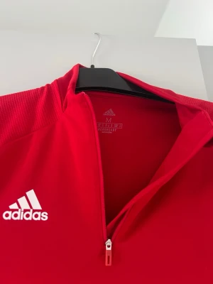 Röd träningsjacka från Adidas - Snygg röd träningsjacka från Adidas med halv dragkedja framtill och vit Adidas-logga på bröstet. Tillverkad i funktionsmaterial med meshpaneler på ärmarna för extra ventilation. Perfekt för träning eller uppvärmning.