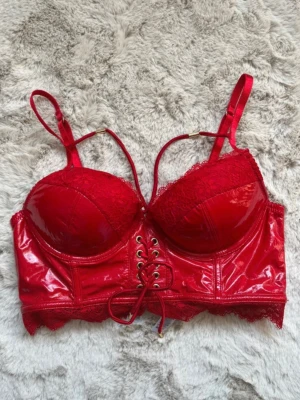 Röd bustier-bh med spets och snörning - Snygg röd bustier-bh med glansigt material och spetsdetaljer. Justerbara axelband, vadderade kupor och snörning framtill för extra attityd. Lång modell med hakknäppning bak och dekorativ spetskant nedtill. Perfekt för dig som vill sticka ut. Ny utan prislapp. 