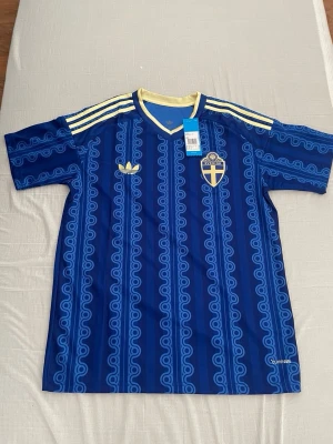 Blå Sverige fotbollströja Adidas - Säljer en blå fotbollströja från Adidas med Sveriges landslagsemblem på bröstet. Tröjan har ljusgula detaljer, klassiska Adidas-ränder på axlarna och ett unikt mönster i blå nyanser. Perfekt för dig som vill stötta Sverige med stil.
