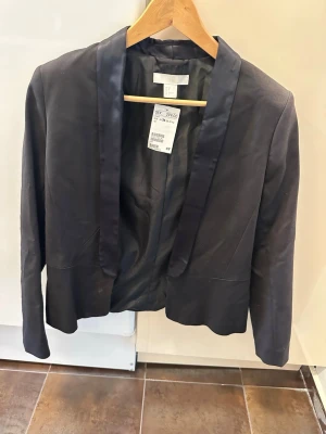 Svart kavaj från H&M, storlek 36 - Stilren svart kavaj från H&M med glansig sjalkrage och öppen front. Kavajen har en klassisk, figurnära passform och är tillverkad i ett slätt tyg som ger en elegant look. Perfekt för att lyfta din outfit med en enkel men snygg touch.
