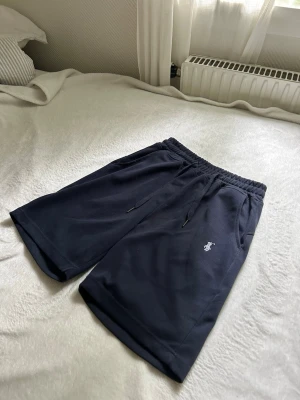 Mörkblå shorts från Polo Ralph Lauren - Säljer ett par mörkblå shorts från Polo Ralph Lauren med klassisk vit logga broderad på vänster ben. Shortsen har dragsko i midjan och två sidofickor. Perfekta för chill dagar eller träning. Sköna och stilrena med avslappnad passform.