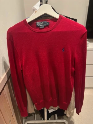 Röd stickad tröja från Polo Ralph Lauren - Röd stickad tröja från Polo Ralph Lauren med rund halsringning och den klassiska blå broderade loggan på bröstet. Tröjan har långa ärmar och ribbade muddar vid ärmslut och nederkant. Perfekt för dig som gillar stilrena och tidlösa plagg.