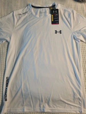 Vit träningströja från Under Armour - Snygg vit funktions-tshirt från Under Armour med svart logga på bröstet och tryck längs sidan. Tillverkad i lätt och snabbtorkande material, perfekt för träning och sport. Rund halsringning och korta ärmar för maximal rörelsefrihet.