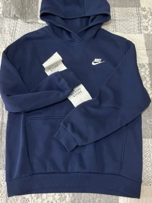 Mörkblå hoodie från Nike - Snygg mörkblå hoodie från Nike med klassisk vit logga broderad på bröstet. Hoodien har huva, känguruficka och ribbade muddar vid ärmar och nederkant. Tillverkad i mjukt bomullsmaterial som känns skönt mot huden. Perfekt för en avslappnad och sportig stil.