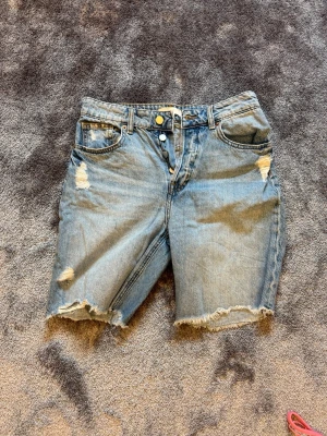 Ljusblå jeansshorts med slitningar - Säljer ett par ljusblå jeansshorts med råa kanter och slitna detaljer framtill. Klassisk femficksmodell med knappgylf och normal passform. Perfekta för en avslappnad stil i sommar.