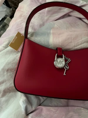 Mörk hallonröd läderväska Michael Kors - Stilren handväska från Michael Kors i mörk hallonröd nyans. Väskan är tillverkad i läder och har en silverfärgad dekor med MK-logga och nyckel. Enkel, rektangulär form med ett stadigt handtag. Perfekt för dig som gillar trendiga accessoarer.