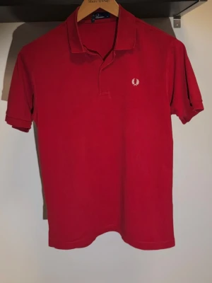 Röd pikétröja från Fred Perry - Klassisk röd pikétröja från Fred Perry med vit broderad logga på bröstet. Tröjan har krage, korta ärmar och knäppning vid halsen. Perfekt för en stilren och sportig look.