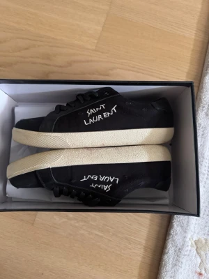 Svarta Saint Laurent sneakers - Svarta sneakers från Saint Laurent med vit broderad logga på sidan. Skorna har en klassisk låg profil, svarta skosnören och vit sula i gummi. Ovandelen är i canvas vilket ger en clean och stilren vibe.