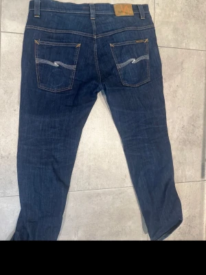 Mörkblå jeans från Nudie Jeans - Snygga mörkblå jeans från Nudie Jeans med klassisk femficksmodell och kontrastsömmar på bakfickorna. Jeansen har rak passform och är tillverkade i ett slitstarkt denimtyg. Perfekta för en avslappnad och trendig look.