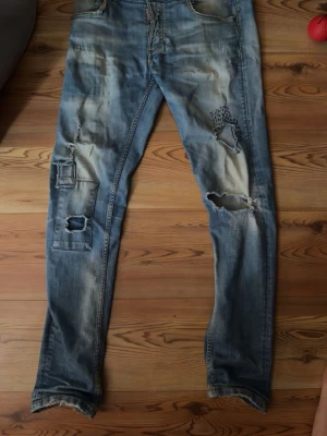 Distressed jeans från Dsquared2 - Säljer ett par blå slitna jeans från Dsquared2 med flera hål och patchade detaljer. Jeansen har en tvättad look, raka ben och klassisk femficksmodell. Perfekta för dig som gillar en edgy och trendig stil.