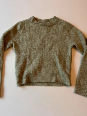 Olivgrön stickad tröja - Mysig olivgrön stickad tröja med rund halsringning och långa ärmar. Tröjan har ribbade muddar vid ärmslut och nederkant samt en rak passform. Perfekt för lager på lager och enkel att matcha med jeans eller kjol.