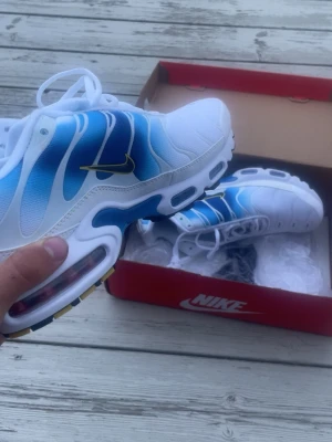 Nike Air Max Plus blå och vita sneakers - Nike Air Max Plus sneakers i vitt och blått med gula detaljer på swoosh-loggan. Skorna har en vågig design på ovandelen, synlig Air Max-dämpning i sulan och vita skosnören. Materialet är en mix av mesh och syntet. Perfekta för dig som gillar sportig och fräsch stil.