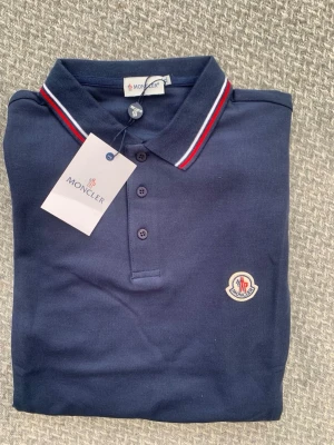 Marinblå polotopp från Moncler - Snygg marinblå polotopp från Moncler med klassisk krage och tre knappar framtill. Kragen har vita och röda ränder som ger en sportig touch. Moncler-logga broderad på bröstet. Materialet är bomull och passformen är normal.