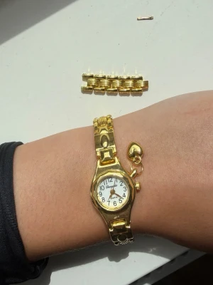 Guldfärgad klocka med hjärthänge - Stilren guldfärgad klocka från Geneva med klassisk rund urtavla och svart text. Armbandet har ett dekorativt hjärthänge och länkar som kan justeras. Perfekt accessoar för dig som gillar detaljer och guldtoner.