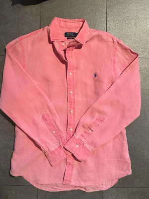 Rosa linneskjorta från Polo Ralph Lauren - Säljer en rosa långärmad skjorta från Polo Ralph Lauren i slim fit. Skjortan är tillverkad i linne och har klassisk krage, knappar framtill och den ikoniska broderade loggan på bröstet. Perfekt för en avslappnad men ändå stilren look.