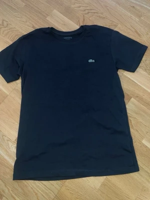 Svart Lacoste t-shirt - Säljer en svart t-shirt från Lacoste med klassisk krokodillogga på bröstet. T-shirten är kortärmad och har rund halsringning. Materialet är mjuk bomull med lite lycra för extra stretch. Perfekt för dig som gillar stilrena plagg med diskret branding.