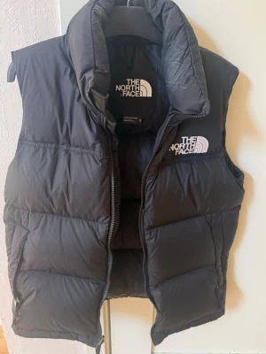 Svart dunväst från The North Face - Snygg svart dunväst från The North Face med hög krage och dragkedja framtill. Västen har broderad logga både fram och bak. Använda cirka 6-7 gånger. Passar inte mig längre.