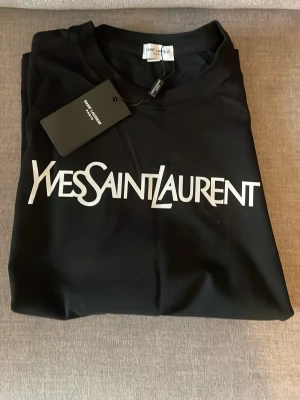 Svart Yves Saint Laurent t-shirt - Snygg svart t-shirt från Yves Saint Laurent med ikoniskt vitt logotryck på bröstet. Klassisk rund halsringning och korta ärmar. Tillverkad i mjuk bomull för en skön känsla och stilren look.