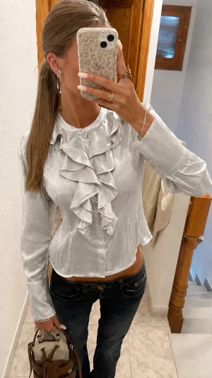  blus med volangdetaljer -  blus med långa ärmar och volangdetaljer framtill. Blusen har en figurnära passform och knäppning framtill. Materialet ser glansigt ut! Den kommer tyvärr inte riktigt till användning 🩶