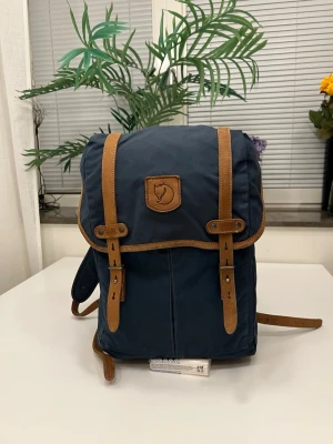 Fjällräven Ryggsäck No.21 Medium i Marinblå - Mörkblå ryggsäck från Fjällräven med snygga bruna läderremmareee och detaljer. Klassisk design med lock och spännen framtill, samt Fjällrävens logotyp på locket. Tillverkad i slitstarkt tyg och har justerbara axelband för extra komfort.                                                            *** Dimensions: 43 cm x 29 cm x 13 cm.  Volume: 20 l.