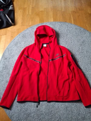 Röd hoodie från Nike med dragkedja - Snygg röd hoodie från Nike med hel dragkedja framtill och svarta detaljer. Hoodien har en huva och två fickor samt en diskret Nike-logga på bröstet. Tillverkad i mjukt material som passar perfekt till streetwear eller chill dagar.