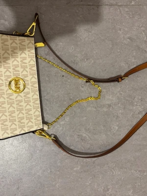 Beige axelväska från Michael Kors - Snygg beige axelväska från Michael Kors med MK-logga i guld framtill. Väskan har ett mönstrat tyg med bruna detaljer, guldfärgade kedjor och justerbar axelrem i brunt läder. Perfekt för dig som gillar stilrena accessoarer med lyxig känsla.