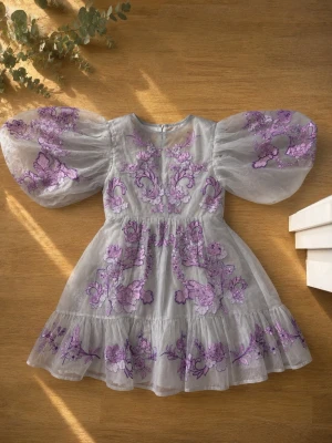 Vit klänning med lila broderi och puffärm - Superfin kort klänning i vit mesh med lila blommigt broderi och stora puffärmar. Klänningen har öppen rygg och volangkant nertill, vilket ger en romantisk vibe. Perfekt för dig som vill sticka ut med en unik och söt look.