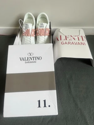 Valentino Garavani vita sneakers - Säljer ett par vita sneakers från Valentino Garavani med ljusrosa band och nitar över ovandelen. Skorna har en chunky sula och är tillverkade i slätt läder med detaljer i gummi. Snygg och modern design som sticker ut med sina ikoniska nitar.