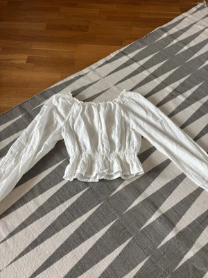 Vit blus med resår och volang - Söt vit blus med långa ärmar, resår i midjan och volangkant nedtill. Blusen har en lätt rynkad struktur och en bred, rundad urringning. Perfekt för en trendig och avslappnad look. Skicka för mer information!💕