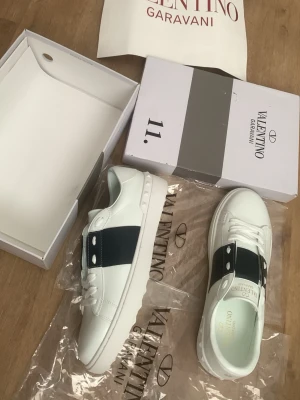 Valentino Garavani vita sneakers storlek 41 - Snygga vita sneakers från Valentino Garavani med bred svart rem över sidan och vita nitar på hälen. Skorna är låga och har en klassisk siluett med vita snören och gummisula. Tillverkade i läder för en exklusiv känsla och look.