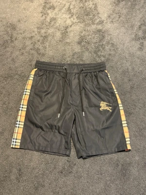Svarta burberrɏ shorts med rutiga sidor - Snygga svarta shorts från Burberrɏ med klassiska beige-rutiga sidopaneler och guldfärgad logga på vänster ben. Shortsen har elastisk midja med snörning och är perfekta för en trendig och avslappnad stil. Passar mer M och pris kan diskuteras