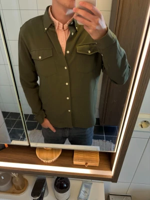 Olivgrön overshirt från Ciszere - Stilren olivgrön overshirt från Ciszere med raka linjer och två bröstfickor med lock och knappar. Skjortan har vita knappar framtill och på fickorna, samt klassisk krage. Perfekt att bära över en t-shirt eller skjorta för en avslappnad look.