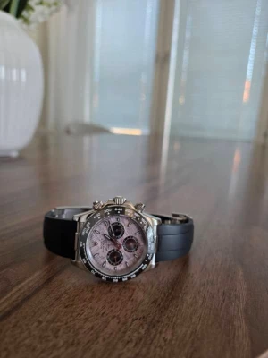 Daytona med svart rubber strap - Säljer en Daytona med svart gummiband och boett i silverfärgad metall. Urtavlan är ljusrosa med marmorerad look och har tre svarta subdials. Klockan har tachymeter på bezeln och klassisk kronografdesign. Använd max 3 gånger så gott som ny, tyärr ingen box medföljer. 