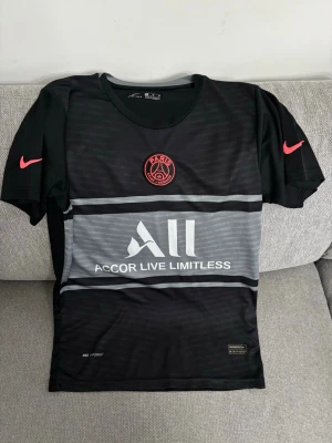 PSG Neymar Jr matchtröja svart - Snygg svart PSG matchtröja från Nike med Neymar Jr och nummer 10 på ryggen. Tröjan har röda detaljer, klubbmärke på bröstet och sponsortryck framtill. Tillverkad i lätt och ventilerande material, perfekt för fotboll eller träning.