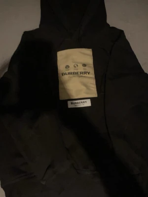 Svart hoodie från Burberry med patch - Säljer en svart hoodie från Burberry med stor beige patch och logga på bröstet. Klassisk huva och ribbade muddar. Hoodien har en modern oversized passform och är tillverkad i mjukt bomullsmaterial. Perfekt för dig som vill ha en streetwear-look.