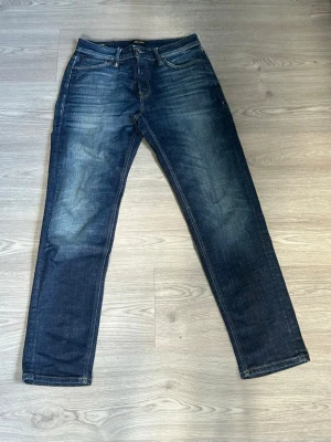 Mörkblå jeans från Jack and Jones  - Snygga mörkblå jeans från Nudie Jeans med klassisk femficksmodell och raka ben. Jeansen har subtila slitningar och kontrastsömmar samt patch i läderimitation bak. Tillverkade i denim med en autentisk tvättad look.