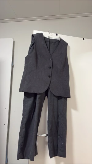 Grå kostym med väst och byxor - Stilren grå kostym med ärmlös väst och matchande raka byxor. Västen har tre knappar framtill och en klassisk v-ringning. Perfekt för dig som vill ha en clean och tidlös look. Materialet känns mjukt och har en lätt struktur. Byxorna är i storlek 40 och västen är i storlek 44